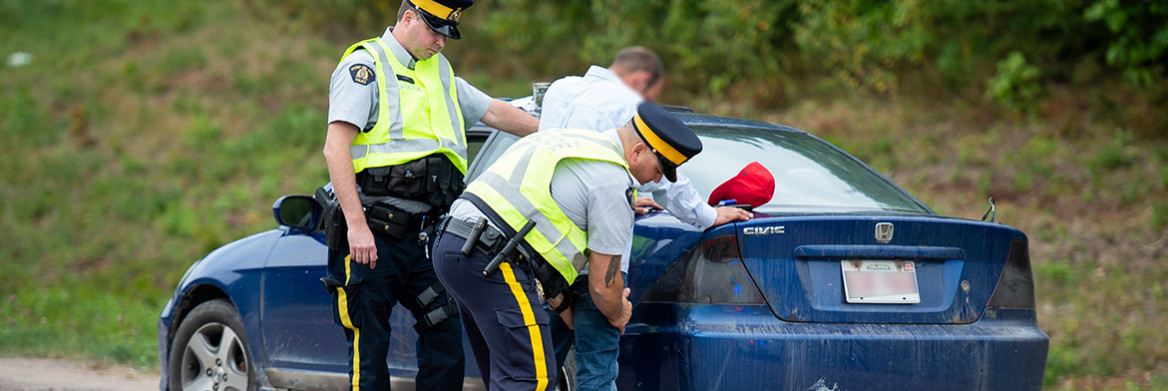 Two RCMP officers inspect a man who leans against a blue car on the side of the highway. 