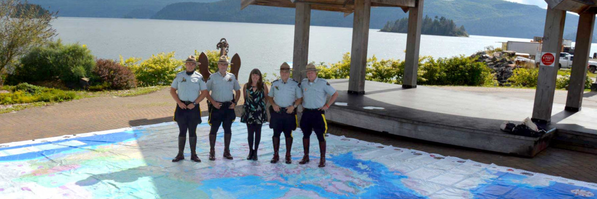 Quatre agents de la GRC et une femme sont debout sur une carte géante du Canada étendue au sol, à l'extérieur, près d'un pavillon. Un lac se trouve derrière eux.